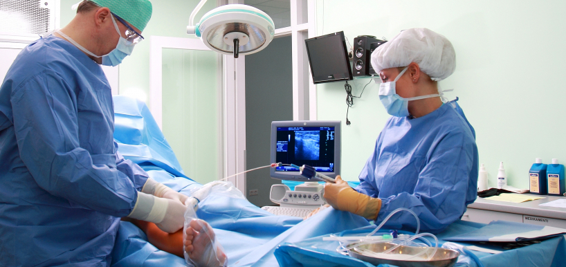 Divi ārsti operāciju zālē veic medicīnisku procedūru pacienta kājai, izmantojot ultrasonogrāfijas iekārtu