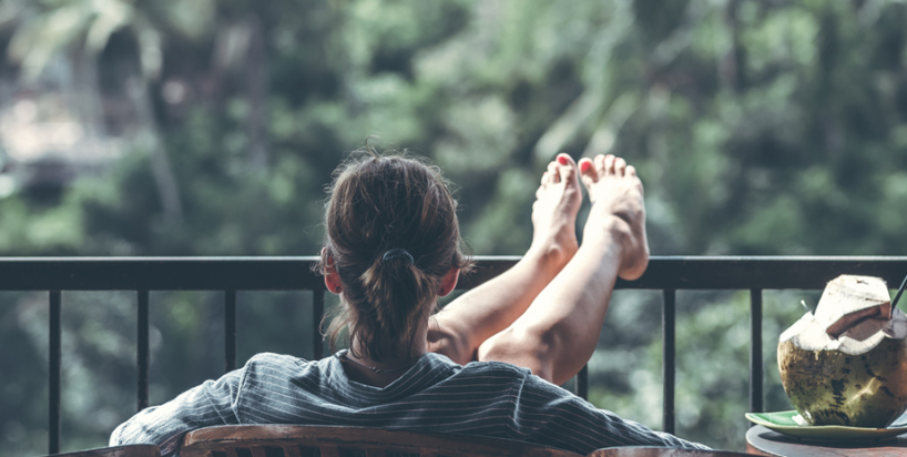 Sieviete atpūšas, sēžot krēslā ar kājām uz balkona margām un skatoties dabā, simbolizējot vieglumu un labsajūtu