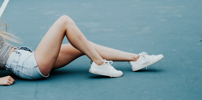 Sieviete sēž ar veselīgām, koptām kājām, valkājot džinsa šortus un baltas kedas uz zilas zemes